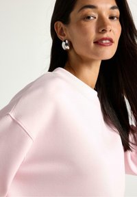 Vaaleanpunainen collegepaita pyöreällä pääntiellä ja lasketuilla harteilla, yhdistettynä kiiltäviin hopeisiin keskiympyräkorvakoruihin, pitkät tummat hiukset omaavalla naisella.