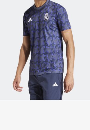 Mies, der ein dunkelblaues Fußballtrikot mit lila geometrischen Mustern sowie den Logos von Real Madrid und Adidas trägt, kombiniert mit navyblauen Adidas-Hosen.
