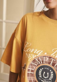 Garinio geltonos spalvos per didelį marškinėlį su balta tekstu ir apvaliu grafiniu motyvu, kuriame yra languotų akcentų, vaizduojančių "Long Island" ir "Hampton."