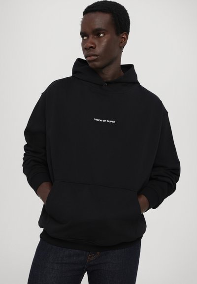Haut à capuche noir en tissu doux, avec une poche kangourou à l'avant et un logo blanc "VISION OF SUPER" sur la poitrine. Coupe simple et décontractée.
