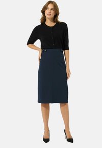 Schwarze Strickjacke mit kurzen Ärmeln und Knöpfen, kombiniert mit einem knielangen marineblauen Bleistiftrock, der Akzentknöpfe am Bund hat.