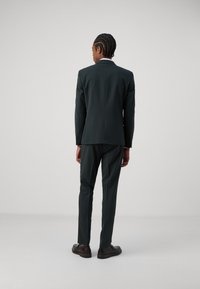 Dark green suit with slim fit, notched lapels, and two-button closure. Worn with brown shoes. Smooth fabric and tailored design.