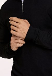 Maglione nero con zip, maniche a costine, texture morbida e zip argentata. Mani che regolano la manica, enfatizzando la vestibilità e il design.