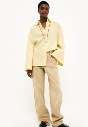 Woman wearing a loose pale yellow buttoned shirt, beige wide-leg pants, black shoes, and a long necklace with a shell pendant.