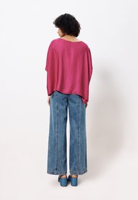 Roze, vloeiende top met een ontspannen pasvorm, gecombineerd met brede blauwe jeans. De outfit heeft open mouwen en is gemaakt van een lichte stof.