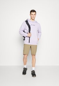 Lila hoodie met voorzak en logo, gecombineerd met kaki shorts. Zwarte rugzak en sneakers. Heldere achtergrond met neutrale tinten.