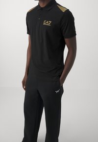 Man in een zwart poloshirt met korte mouwen, voorzien van het gouden EA7 Emporio Armani-logo en gestreepte schouderdetails, gecombineerd met zwarte broek met wit logo.