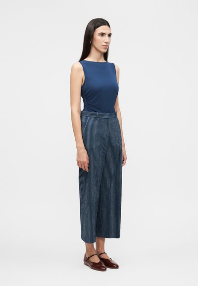 Γυναίκα με μακριά σκούρα μαλλιά φοράει αμάνικη μπλε μπλούζα, μπλε φαρδιά, καρό cropped παντελόνια και καφέ παπούτσια Mary Jane, στέκεται πλαγίως.