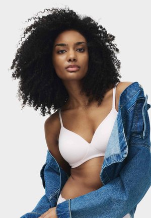 Frau mit voluminösem lockigem Haar, die einen blassrosa BH und eine blaue Jeansjacke trägt, die leicht von den Schultern rutscht, vor einem schlichten Hintergrund.