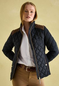 Veste matelassée bleu marine avec accents en daim brun sur les épaules, garnitures dorées et logo brodé. Associée à une chemise blanche et une ceinture marron.