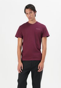 Burgunderfarget bomulls T-skjorte med klassisk rund hals og korte ermer. Har et lite hvitt "TOMMY JEANS"-logo på venstre bryst.