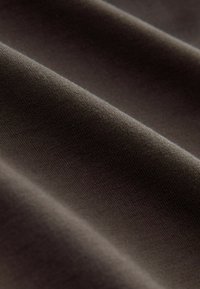 Hnědá látka s mírně texturovaným povrchem, vykazující jemné, skládané záhyby a detaily jemného šití. Lehký a pružný materiál.
