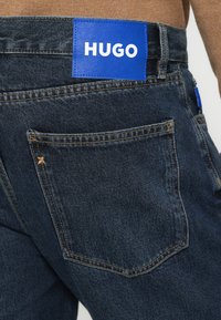 Sötétkék farmernadrág hátsó zsebbel, látható varrással és a derékrész közelében elhelyezett, "HUGO" feliratú kék téglalap alakú címkével.