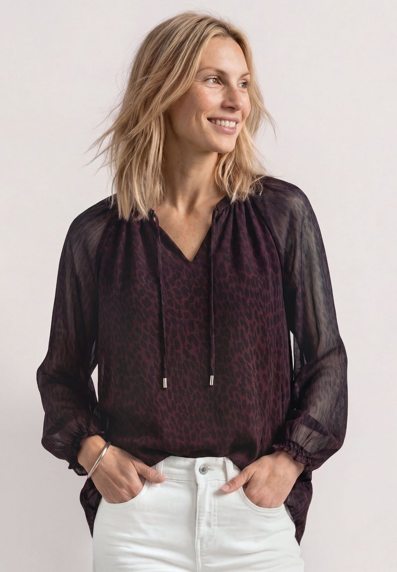 Femme aux cheveux blonds mi-longs portant un chemisier violet transparent à motifs et un pantalon blanc, les mains dans les poches, souriante et regardant vers la droite.