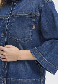 Denim košulja tamnoplave boje s dva džepa na prsima, dugmadi na preklopu i širokim rukavima. Sadrži kontrastne šavove i malu oznaku branda.