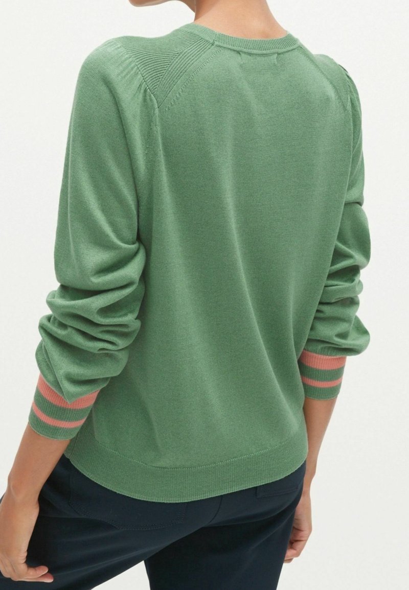 Femme portant un pull vert à manches longues avec des poignets rayés rose et vert, associée à un pantalon foncé, vue de dos.