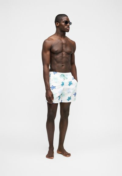 Μυώδης άνδρας φορώντας γυαλιά ηλίου και λευκό μαγιό με μπλε και πράσινα σχέδια χελωνών, όρθιος ξυπόλητος μπροστά σε λευκό φόντο.