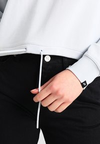 Světle modrá mikina se stahovací šňůrkou na spodním lemu a žebrovanými manžetami, spárovaná s černými kalhotami. Obsahuje stříbrný knoflík a značkovou visačku.