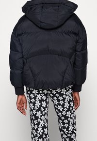 Černá péřová bunda s kapucí, která má prošívaný design a elastické manžety, v kombinaci s kalhotami s květinovým vzorem v černé a bílé barvě.