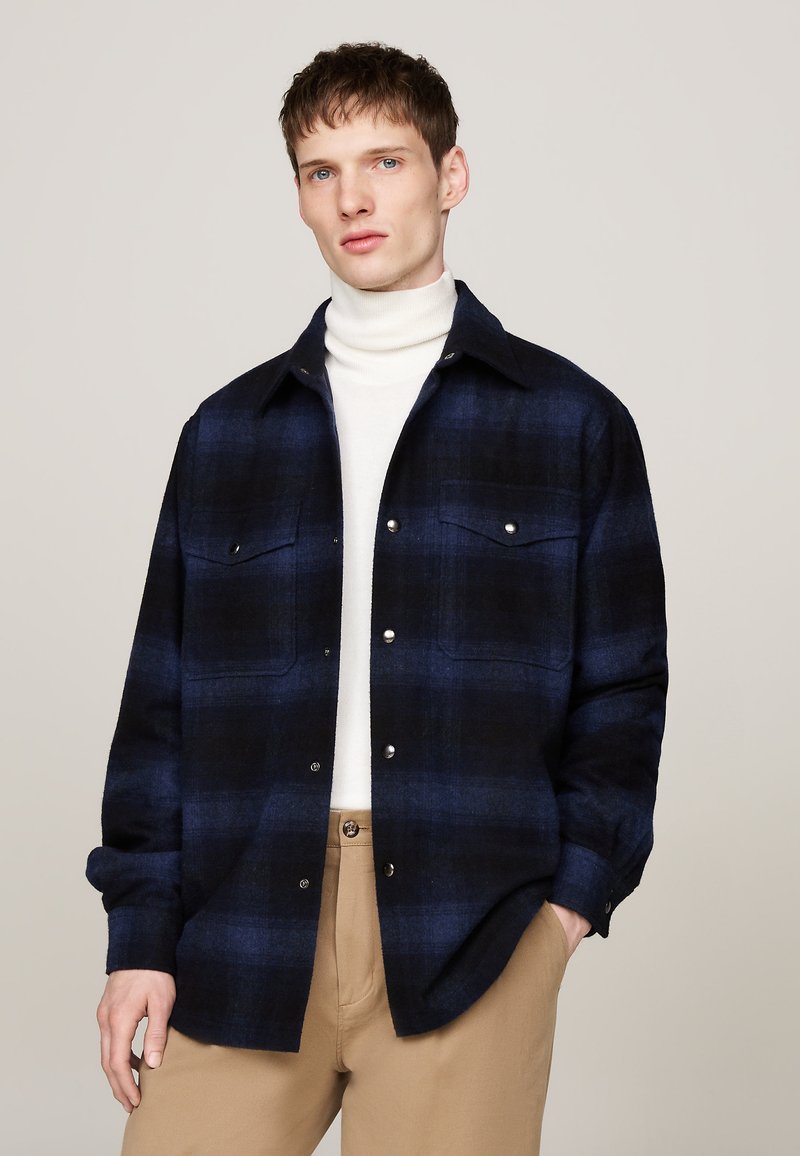 Navy and black plaid flannel shirt with button-up front, two chest pockets, snap buttons, and soft texture, paired with a white turtleneck.