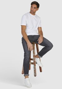 Biała koszulka z krótkim rękawem, ciemnoszare dżinsy i białe trampki. Siedząc na drewnianym stołku, który jest widocznie zużyty i ma postarzany wygląd.