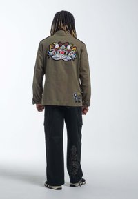 Khaki kurtka wojskowa z kolorowym haftem orła na plecach, połączona z czarnymi spodniami cargo z ozdobnym wzorem.