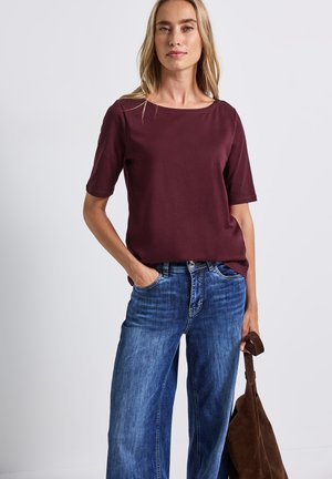 Top bordeaux à manches courtes avec un col bateau associé à un jean en denim bleu large. Le modèle tient un sac en daim marron. Texture de tissu lisse.