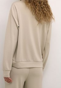 Sweat-shirt beige à manches longues avec poignets côtelés, doté d'une coupe décontractée et d'une texture lisse, accompagné d'un pantalon assorti.