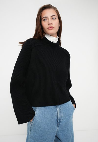 Black ribbed knit sweater with a high neck and dropped shoulders, paired with light blue denim pants. Smooth texture, loose fit.