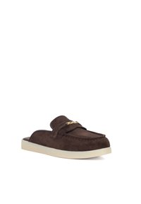 Bruine suède instappers met een gouden kettingaccent, met een ronde neus en een witte rubberen zool voor contrast.