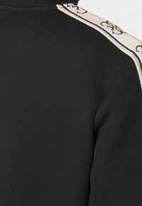 Prenda de tela negra con ribete de color beige claro que presenta un patrón repetido de logotipos negros entrelazados a lo largo de la costura del hombro.