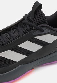 Gros plan d'une chaussure de sport noire avec tige en mesh, lacets noirs, bandes diagonales blanches et semelle grise avec accents violets et orange.