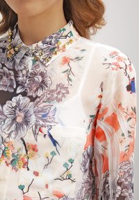 Blouse en voile floral arborant un design multicolore avec des fleurs violettes, oranges et roses ; elle possède un col clouté et une poche sur le devant.