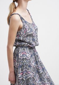 Młoda kobieta z blond kucykiem, ubrana w bezrękawnikową sukienkę w wzory paisley w odcieniach niebieskiego, białego i różowego, na jednolitym tle.