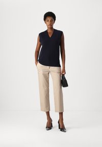 Maglione senza maniche blu navy con scollatura a V, pantaloni cropped crema e décolleté in vernice nera. Il modello tiene una piccola borsa nera.