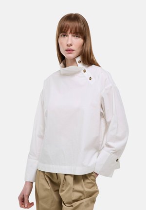 Femme aux cheveux châtain clair portant un chemisier blanc à col haut avec des boutons et un pantalon beige ample, debout devant un fond uni.