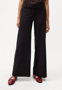 Pantalons larges noirs avec une texture de tissu lisse. Présente un devant plat, des passants de ceinture et tombe lâchement jusqu'à la cheville. Associé à des chaussures plates rouges.