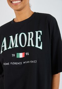 Juodas medvilninis marškinėliai su užrašu "AMORE" žaliai, Italijos vėliavos grafika viduryje, ir tekstu "ROMA FLORENCIJA MILANAS VENECIJA" apačioje.