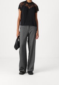 Czarny top z koronkową lamówką, w zestawieniu z szarymi spodniami o szerokich nogawkach. Modelka trzyma małą czarną torebkę. Tkanina wygląda na gładką i strukturalną.