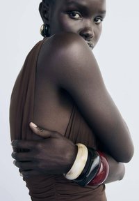 Robe marron, texture lisse, design ajusté. Quatre bracelets : un blanc, un noir, un rouge, un marbré. Peau noire, bras croisés.