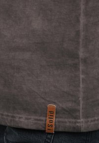 Grå bomulds t-shirt med en blød tekstur, der har en synlig sy-seam og et brunt lædermærke med mærket "Solid" nær kanten.