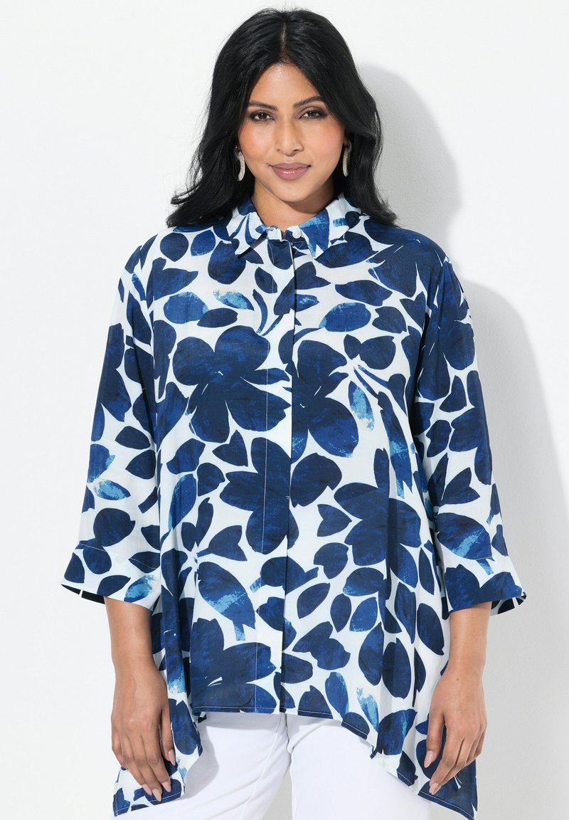 Camicia a maniche tre quarti di colore blu con motivo floreale, dal taglio morbido e con colletto, caratterizzata da un orlo asimmetrico su uno sfondo chiaro.