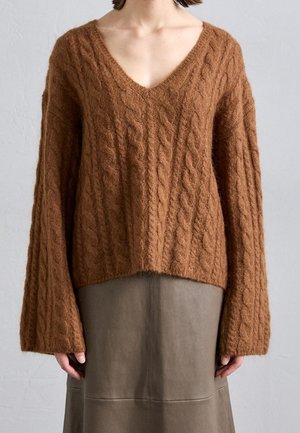 Femme portant un pull marron à col en V en maille torsadée avec des manches longues et une jupe de couleur taupe sur un fond uni.