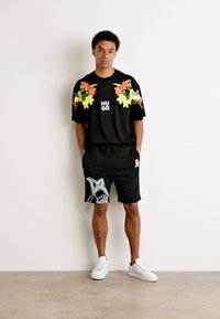Chemise noire à manches courtes avec un design floral, logo "HUGO". Shorts noirs avec art abstrait et taille élastique. Baskets blanches.