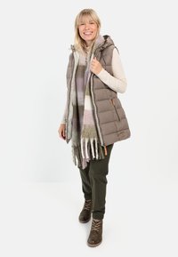 Polstret, ærmeløs vest i taupe med hætte og lynlås. Den har en orange detalje og ledsages af et stort tørklæde med ternet mønster i bløde farver.