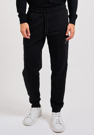 Hombre vestido con pantalones jogger negros con cordón y zapatillas blancas, de pie frente a un fondo liso.