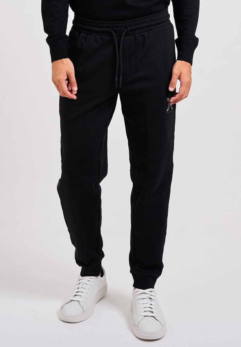 Hombre vestido con pantalones jogger negros con cordón y zapatillas blancas, de pie frente a un fondo liso.