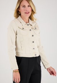 Vrouw in een beige denimjack met knoopdetails, zwarte broek, gouden creolen en een gouden armband, poseert tegen een effen achtergrond.