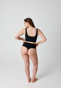 Vrouw staat op blote voeten, met haar rug naar de camera, terwijl ze een zwarte naadloze ondergoedset rechtzet tegen een effen lichte achtergrond.