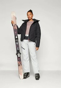 Čierna prešívaná bunda, sivé lyžiarske nohavice a čierne lyžiarske topánky. Drží lyže s gradientovým dizajnom. Obsahuje červený akcent. Vertikálny silueta.